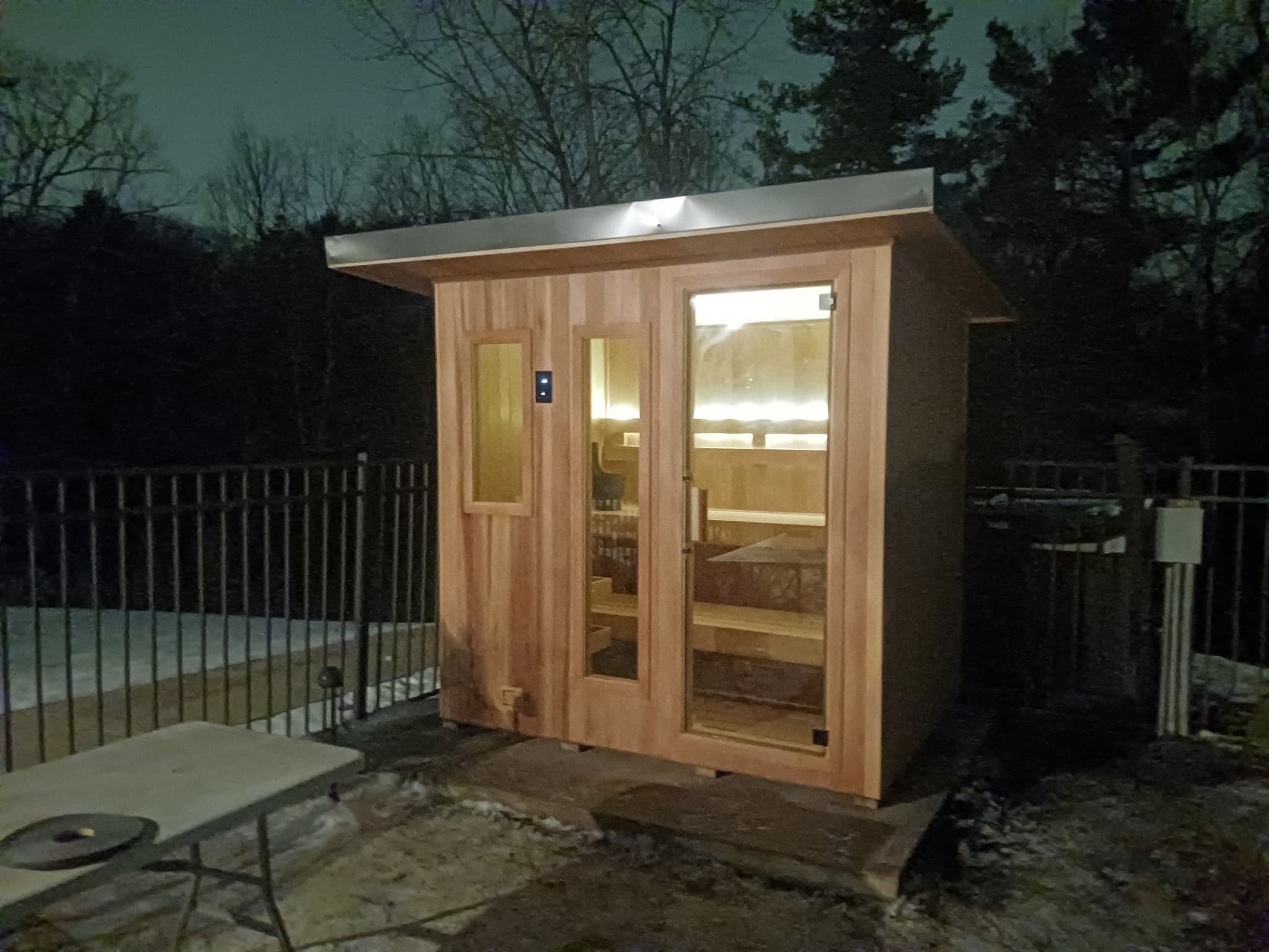Outdoor sauna cabin lit at dusk beside a pool.