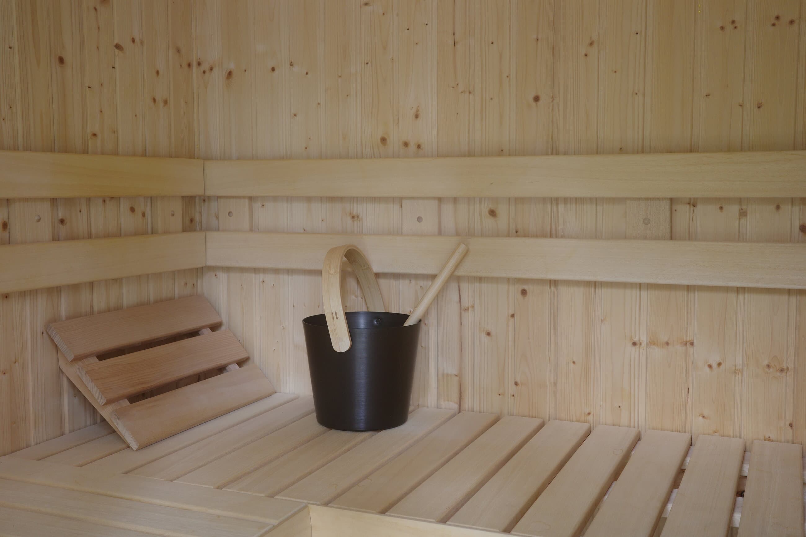 Finished pale wood sauna bench with black bucket, ladle, and backrest detail.