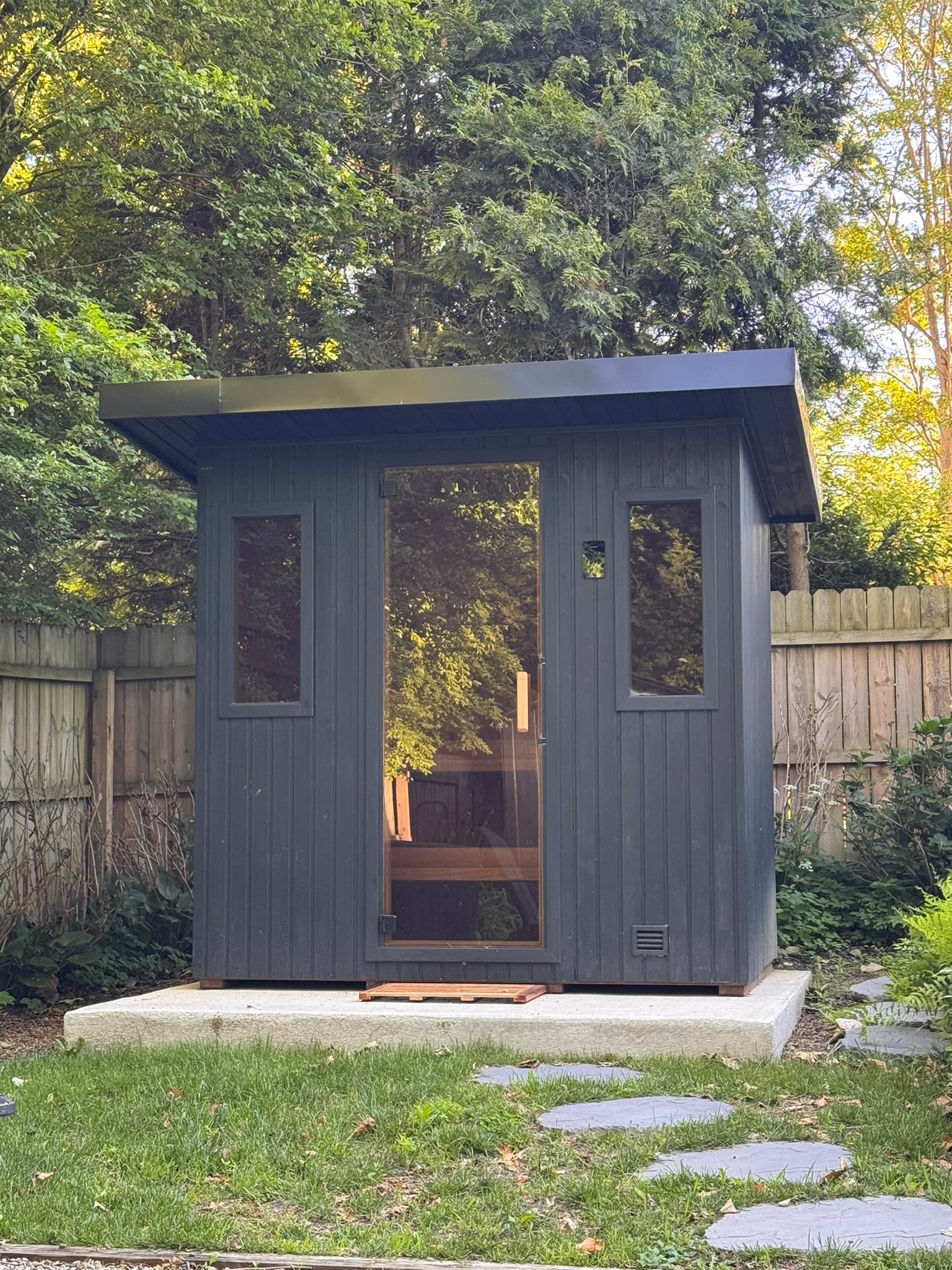 Dark freestanding outdoor sauna with a glass door in a landscaped backyard.